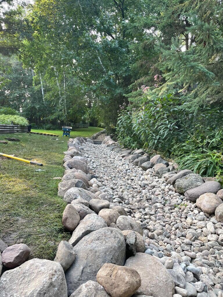 A long boulder-lined rip rap drainage channel curving through a wooded backyard, carrying water safely past mature trees and plantings